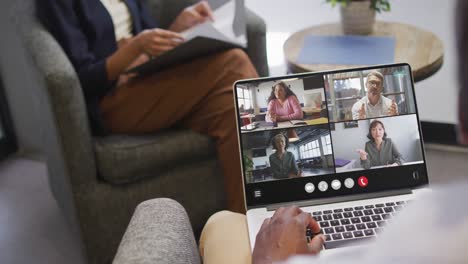hombre afroamericano usando una computadora portátil para una llamada de video, con diversos colegas de negocios en la pantalla