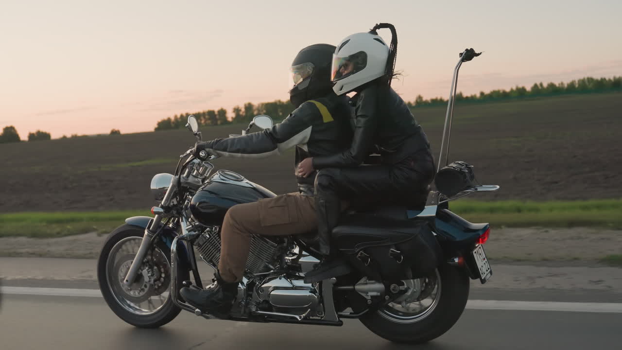 Young adult couple wearing helmets ride motorcycle on countryside road during sunset, passenger embracing rider as they travel across vast open fields