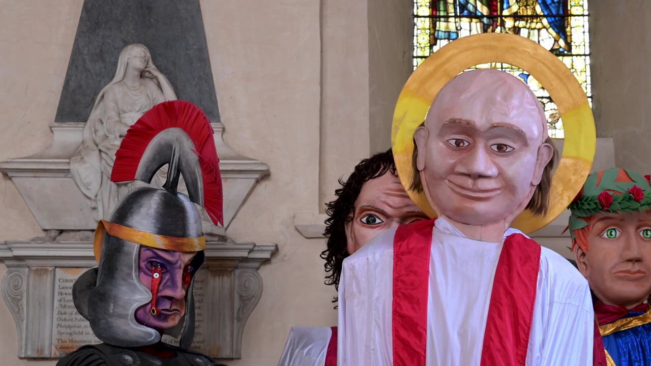 Grandes cabeças de fantoches pintadas e modelos sagrados estão em frente a um monumento de pedra e vitrais dentro da Catedral de St Albans.