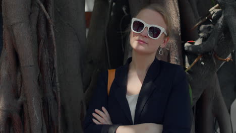 a woman in sunglasses and a blazer stands in front of a tree with roots