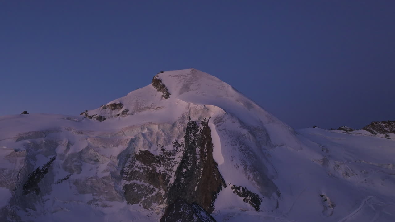 A breathtaking aerial view of the Allalinhorn moments before sunrise. Soft dawn light paints the snow-covered summit and surrounding Swiss Alps in warm pastel hues