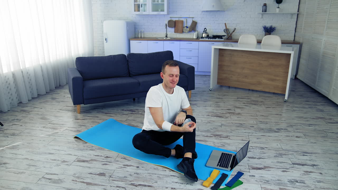 Young man exercising at home. Man is sitting on a mat on floor and holding balance with a bottle. Fitness at home during quarantine.