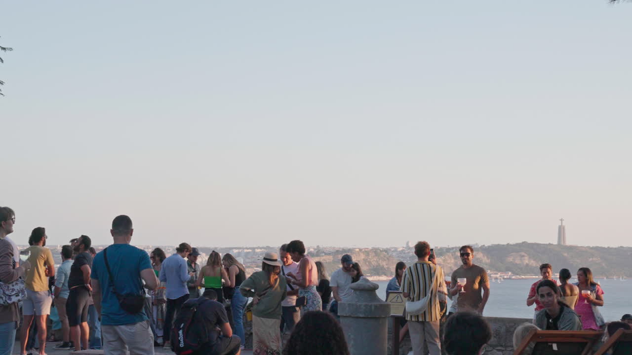 A crowd of tourists stands atop a viewpoint in Lisbon, Portugal, gazing out over the cityscape during a mesmerizing sunset, with the Tagus River glowing in the distance.