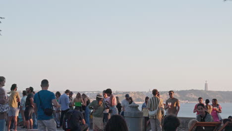a crowd of tourists stands atop a viewpoint in lisbon, portugal, gazing out over the cityscape during a mesmerizing sunset, with the tagus river glowing in the distance.