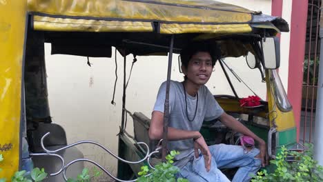 an young indian auto driver in grey t-shirt sitting inside the auto and looking at the camera