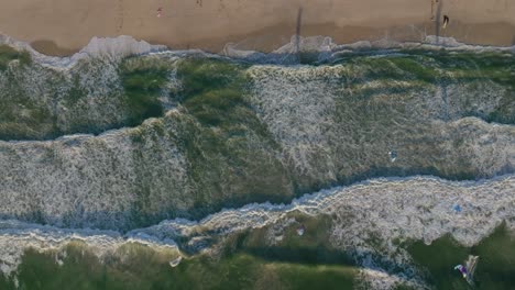 slow motion top-down drone footage shows atlantic ocean waves rolling and foaming as they crash on blouberg beach near cape town, with surfers scattered near the sandy shoreline and entering water