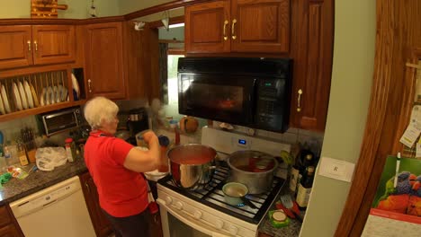 mujer mayor de pie en la cocina en la estufa mientras echa jugo de tomate fresco en tarros de conservas