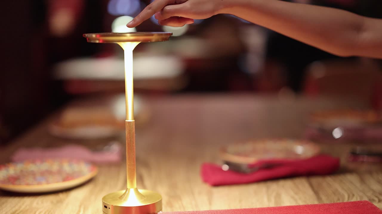 A hand taps the top of a modern touch lamp, illuminating a wooden restaurant table set with plates and napkins in warm ambient lighting