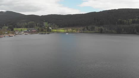 wide panoramic view of a calm lake with buildings at the shore surrounded by forest and hills, as a boat sailing through the water