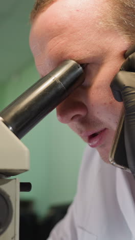 una vista de cerca de un hombre con una bata blanca de laboratorio y guantes negros, hablando por teléfono mientras observa de cerca a través de un microscopio en un entorno de laboratorio