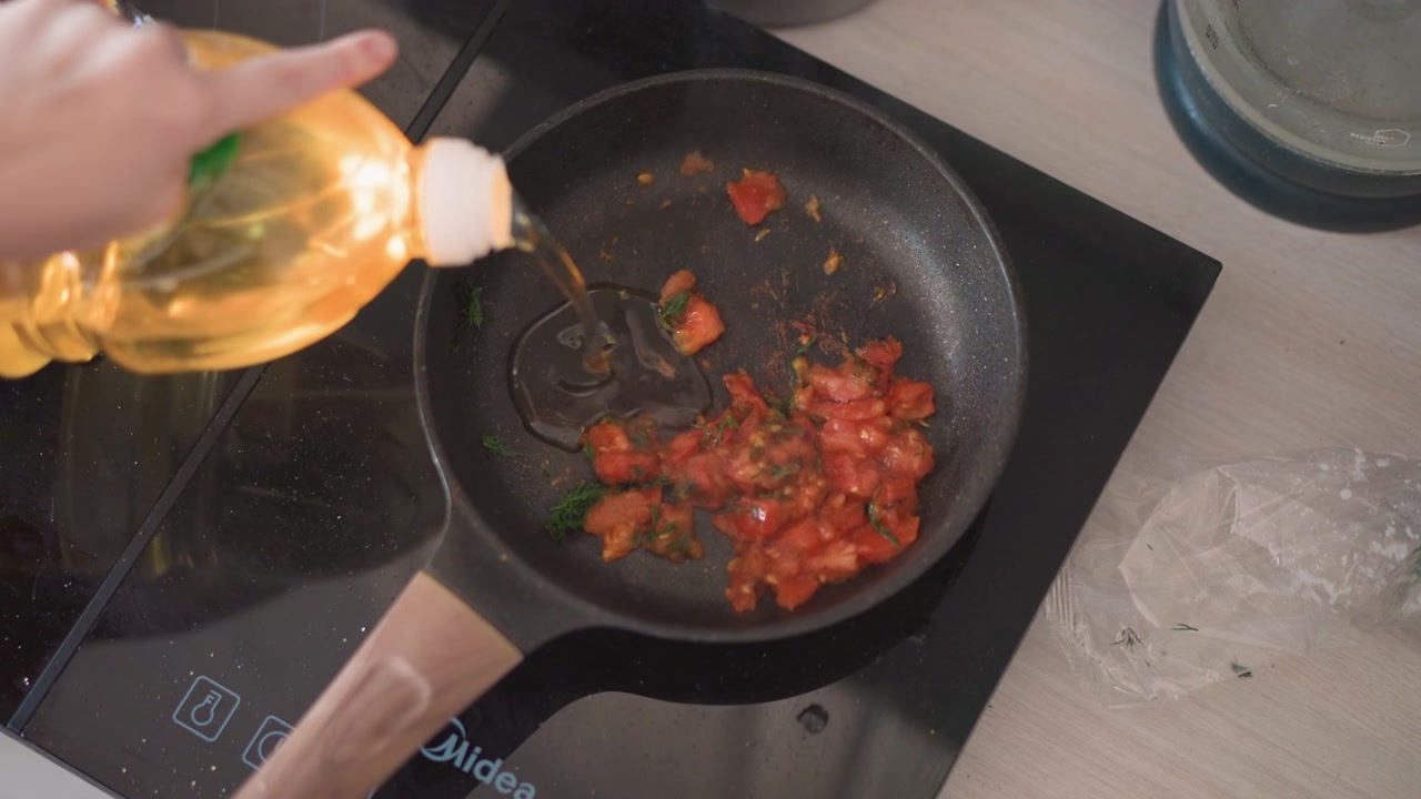 Momento di cucina con olio d'oliva. Blogger che versa olio d'oliva pregiato in un piatto di pomodoro caldo sul bancone. Primo piano dell'olio che cola nella padella di pomodoro mentre il blogger versa olio d'oliva dalla bottiglia alla luce del mattino.