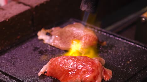 detailed view of meat pieces being seared with a torch on a hot grill surface.