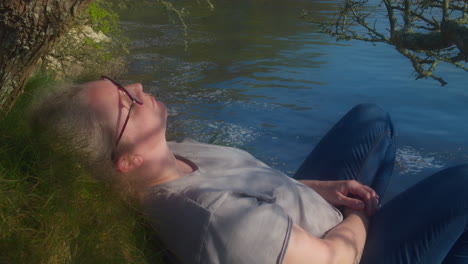 woman in glasses relaxing with eyes closed lying on a grassy hillside next to a lake on a beautiful sunny day