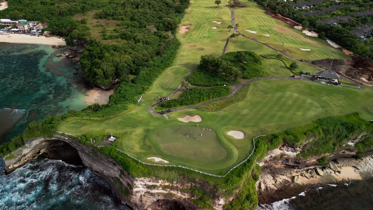 Aerial drone footage of a coastal Bali golf resort with players on ocean cliff fairways large putting greens sand bunkers paved resort roads and waves breaking along the rocky shoreline below