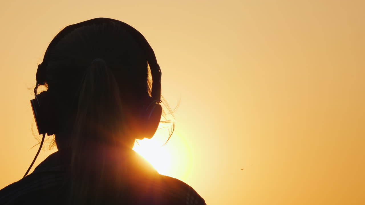 силуэт девушки в наушниках, слушающей музыку на фоне большого заходящего солнца ан