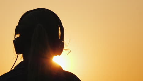 Silhouette-Of-A-Girl-In-Headphones-Listening-To-Música-Against-The-Backdrop-Of-A-Large-Setting-Sun-An