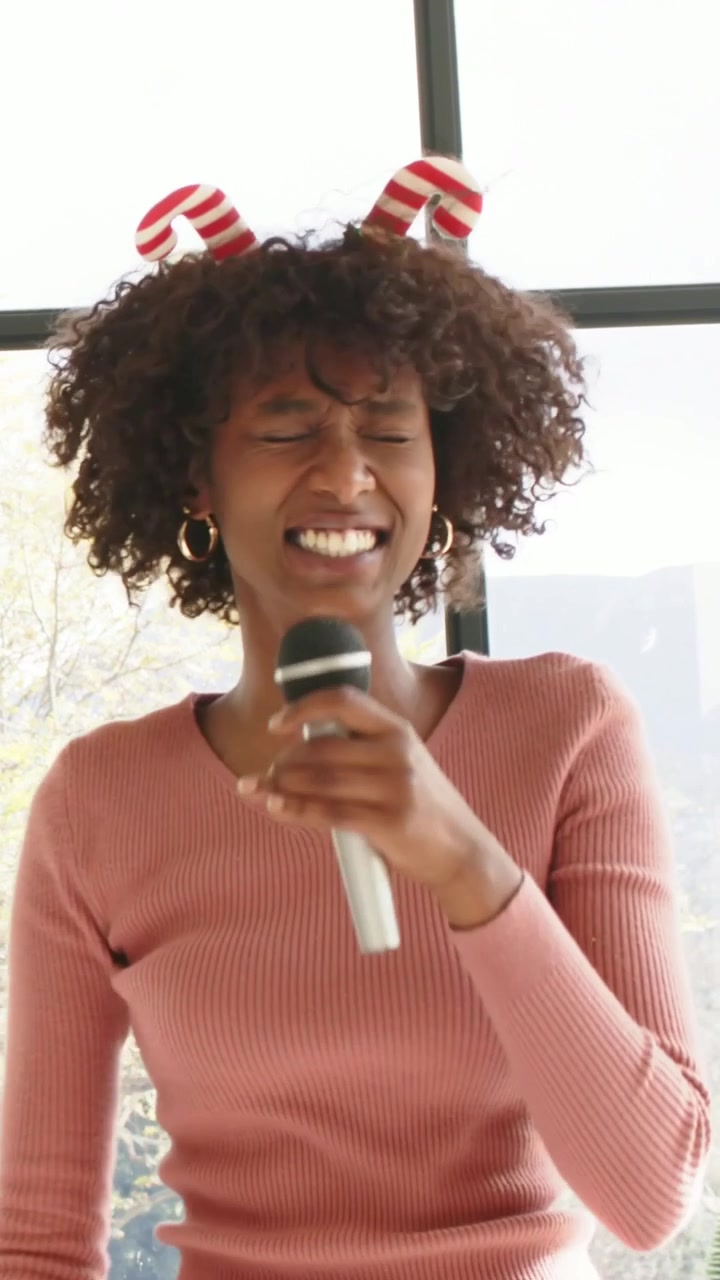 Vertical video: Singing woman holding mic to mouth and adjusting red headband in pink top at window