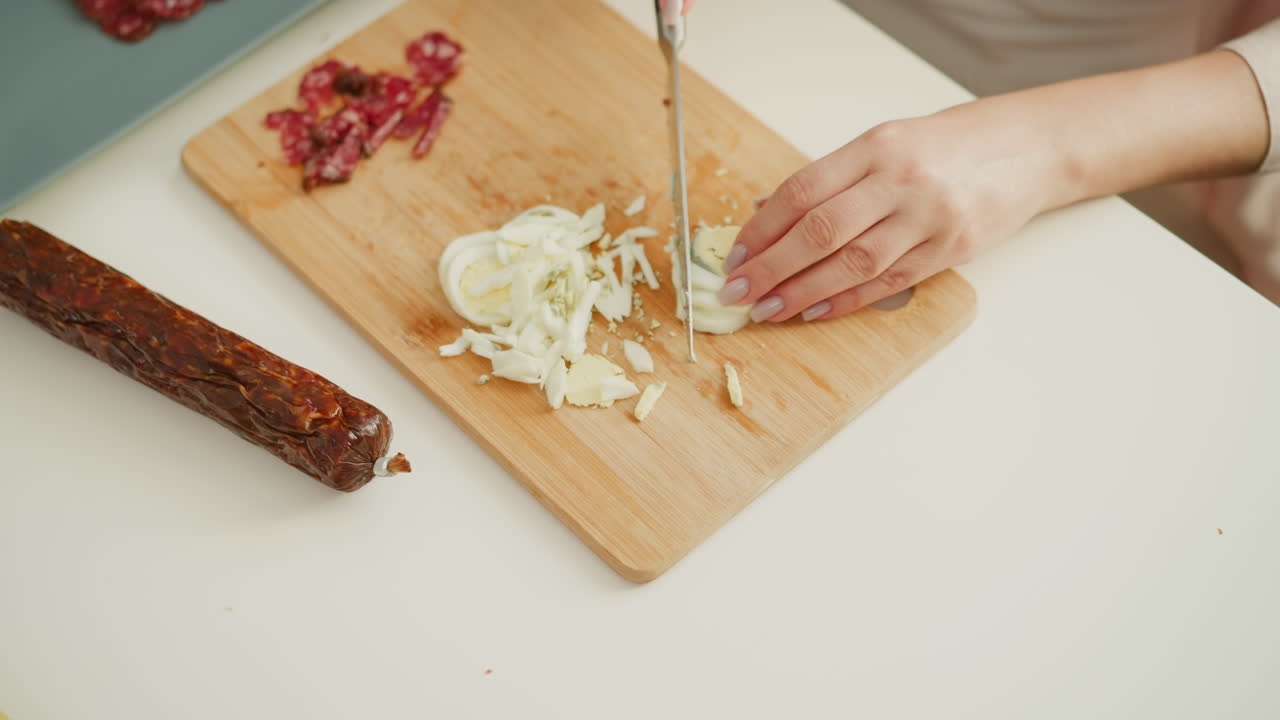 Hand of head chef slicing boiled egg into tiny pieces beside sliced sausage on table, using precise knife skills on wooden cutting board in bright kitchen setting, focusing on culinary preparation
