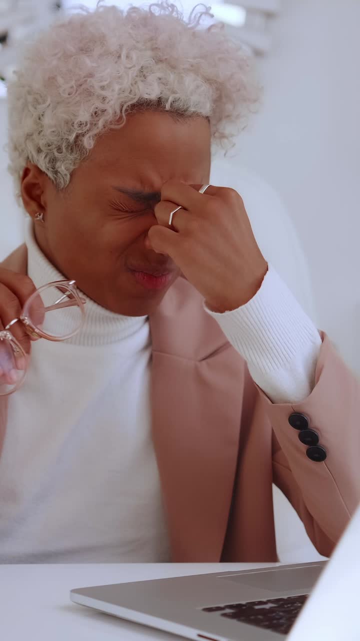 Young woman rubbing eyes holding eyeglasses while seated at a desk with laptop