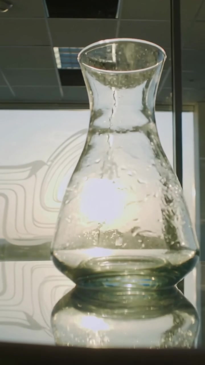 Vertical video: Sunlight brightening glass carafe showing droplets and mirror reflection in office
