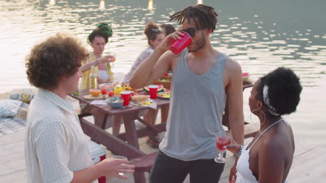 multiethnic friends chatting over drinks at party on lake pier