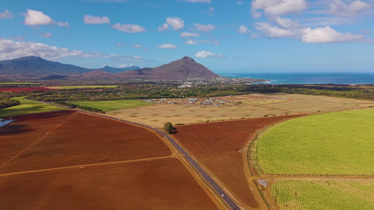 Wide aerial shot of a road on Mauritius island landscape with coastal lagoons, towns, and distant mountains under dramatic tropical clouds and sunlight