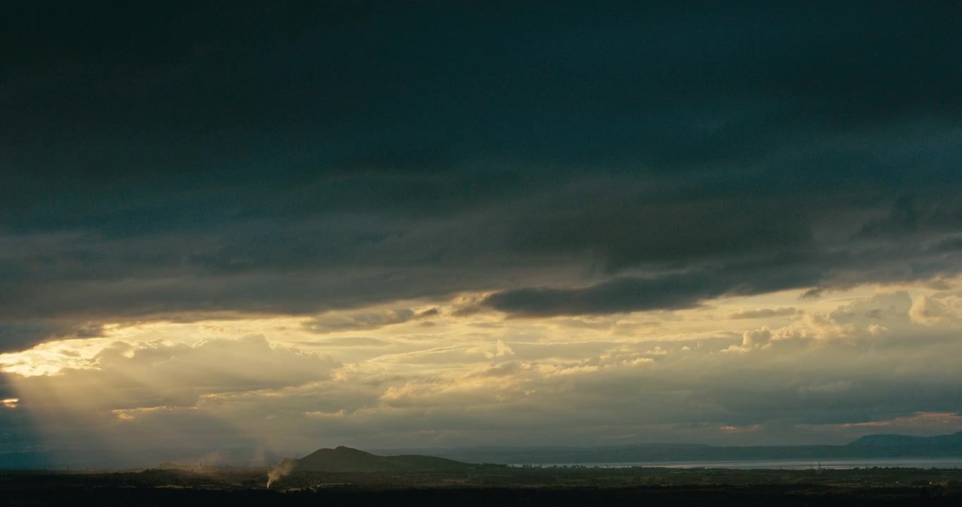 Dark storm clouds with dramatic sunbeams breaking through over a distant landscape. Moody weather, layered clouds, and atmospheric light create a powerful natural panorama.