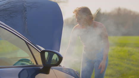 woman broken down on country road looking under smoking bonnet of car