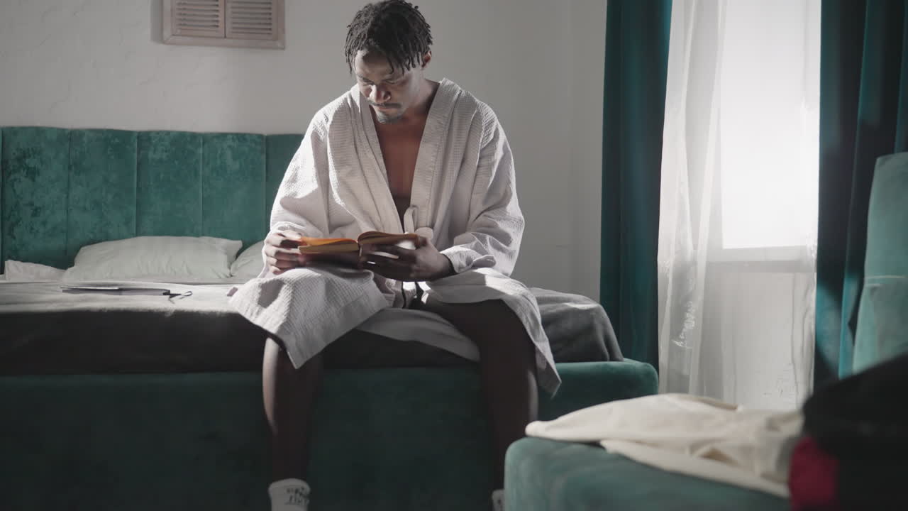 Man in white bathrobe sitting on bed, reading book in morning light. Comfortable, quiet indoor setting with backpack on bed, socks and sandals worn. Tranquil, focused atmosphere