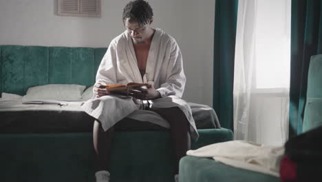 man in white bathrobe sitting on bed, reading book in morning light. comfortable, quiet indoor setting with backpack on bed, socks and sandals worn. tranquil, focused atmosphere