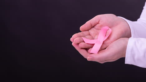 Video-of-midsection-of-caucasian-woman-holding-pink-cancer-awareness-ribbon,-with-black-background