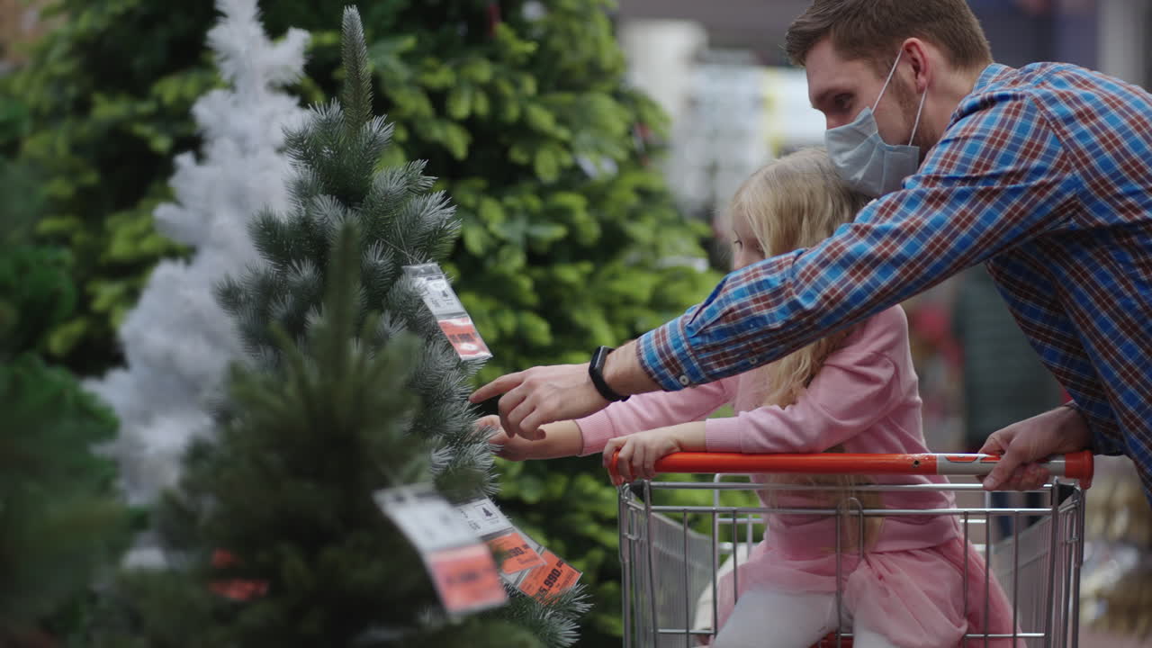 papá e hijo eligen juguetes de navidad en el estante de la tienda. padre e hija pequeña en el carrito de compras eligen decoraciones de navidad. cámara lenta en el centro comercial