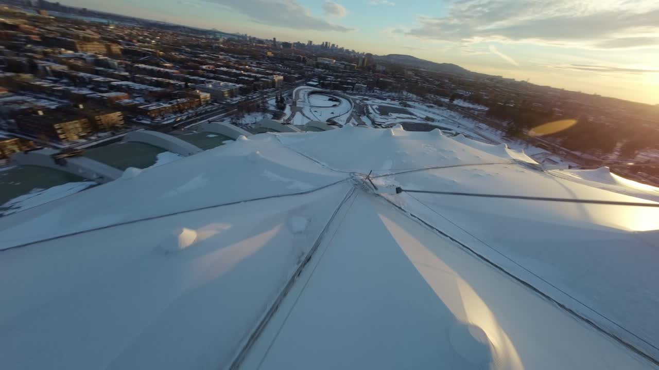 Drone swooping above Montreal Olympic Stadium, snowy winter day, with sunset casting warm light