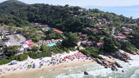 escena de drones aéreos de barrio residencial frente a la playa con mucha naturaleza arena mar y costas rocosas con mucha gente divirtiéndose en la playa muchas sombrillas en verano florianópolis jurere