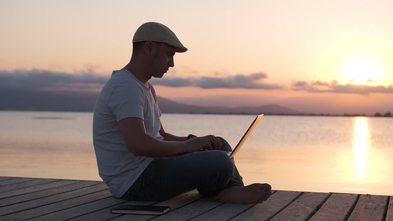 Male traveler sitting on the shore using laptop. Digital nomad or remote worker concept