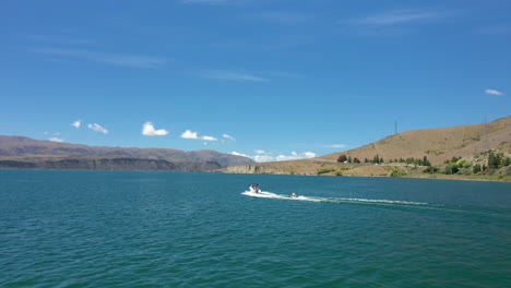 a drone shot of man and a boy being towed on an inflatable tube behind a speed boat zig-zagging in and out of the wake on the beautiful blue lake aviemore in new zealand