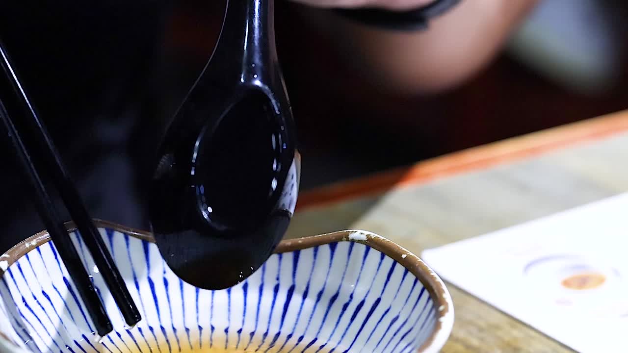 A person carefully holds a black spoon above a blue-striped bowl on a table.