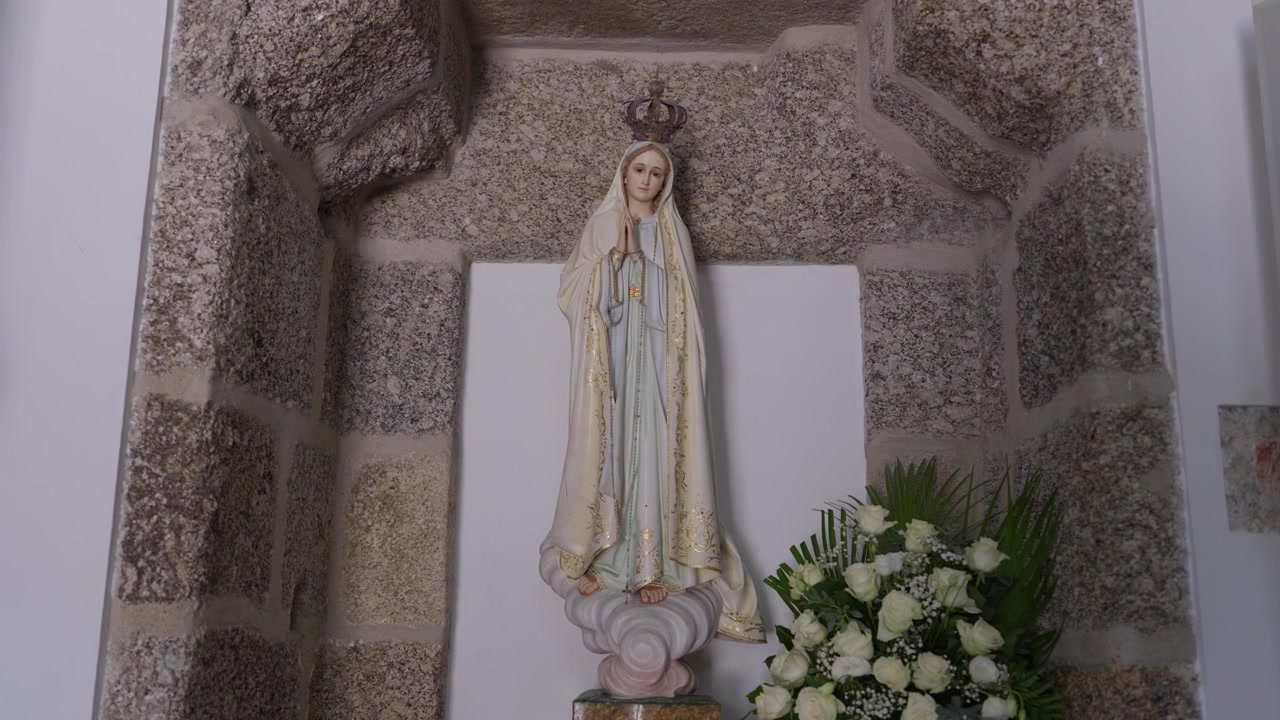 Estátua da Virgem Maria coroada e com manto, em pé em um nicho de pedra ao lado de rosas brancas