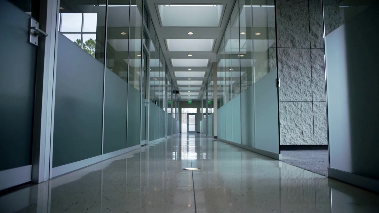Cámara baja avanzando por un pasillo de oficina, revelando particiones de vidrio y una salida