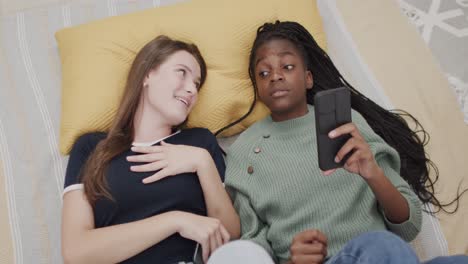 happy diverse teenage female friends lying on bed using smartphones, talking and laughing