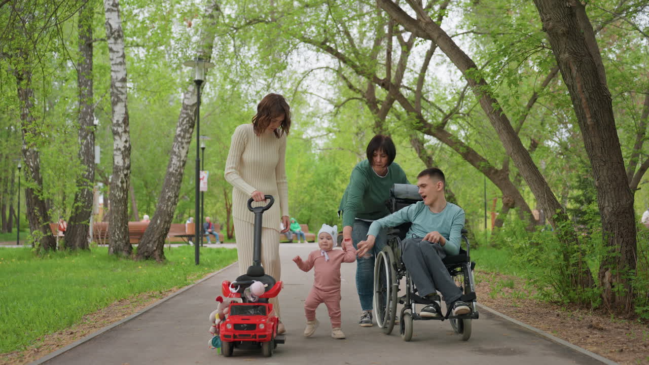 Walking Together With Stroller And Wheelchairs Outdoors, Cheerful Family Enjoying Sunny Day In Nature, Group Of Family Members Exploring Treelined Pathway With Mobility Aids And Children