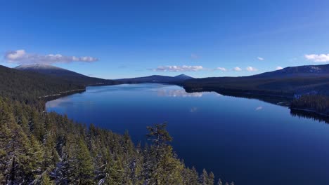 Drone-through-snowy-forest-trees-with-reflective-blue-lake-sunny,-60fps