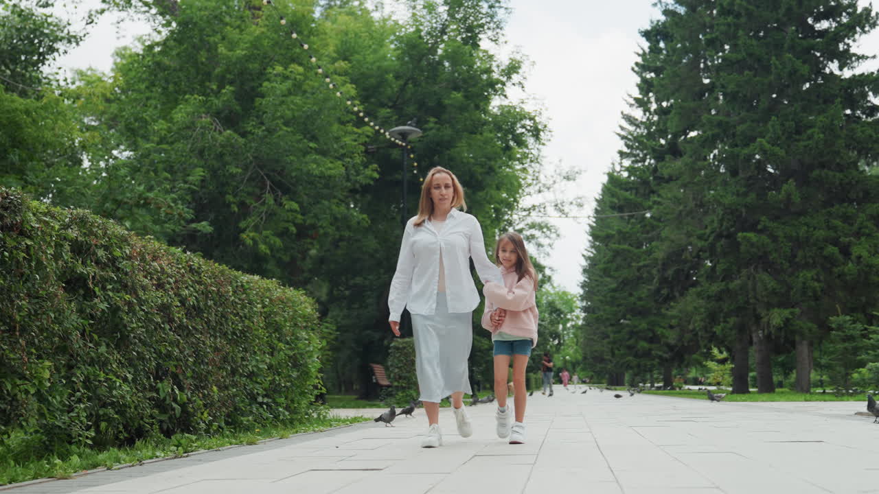Moeder en dochter wandelen in het park langs een pad omzoomd met heggen, casual kleding, moeder loopt iets voorop terwijl kind dichtbij volgt, verhard pad, hoge bomen, sereen daglicht, spontaan familiemoment, ontspannen