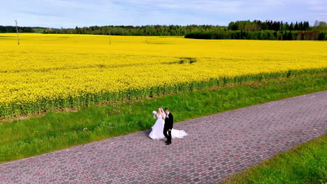 a newlywed couple walks along a cobblestone road bordering a bright canola field as a smooth low drone tracks beside them, capturing joyful celebration, movement, and pastoral romance