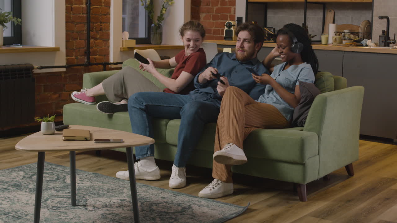 tres compañeros de habitación usando un teléfono inteligente, jugando videojuegos y escuchando música con un teléfono inteligente sentado en un sofá en casa 2