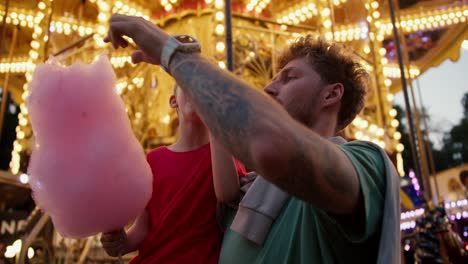 un niño rubio con una camiseta roja come azúcar de algodón rosa con su padre en una camisetta verde contra el telón de fondo de una hermosa atracción brillante con flores amarillas en un parque de atracciones por la noche