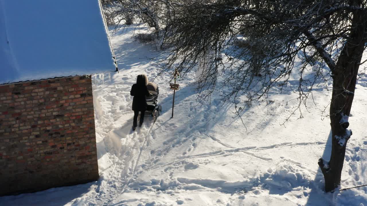 아기 마차와 함께 산책하는 어머니는 나무와 함께 눈 인 정원에 오두막 벽돌 벽을 통과합니다.