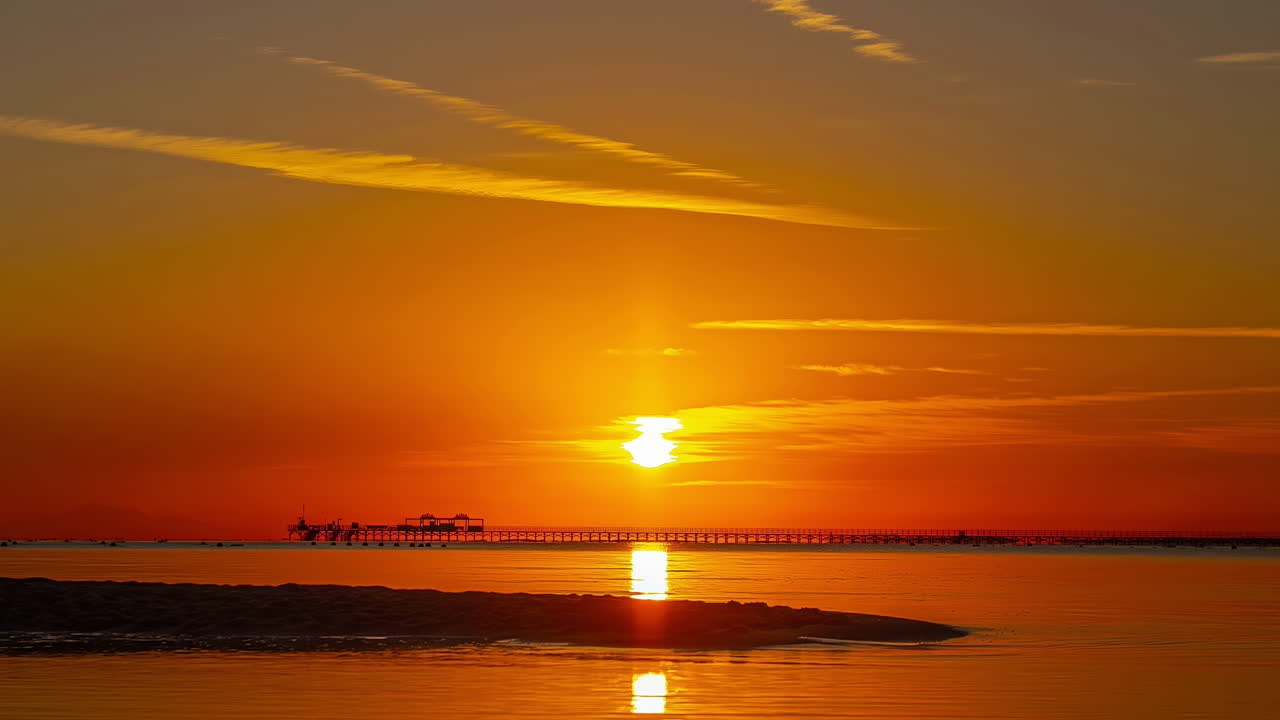 Sunset over water with vibrant orange skies and silhouette of a pier, timelapse