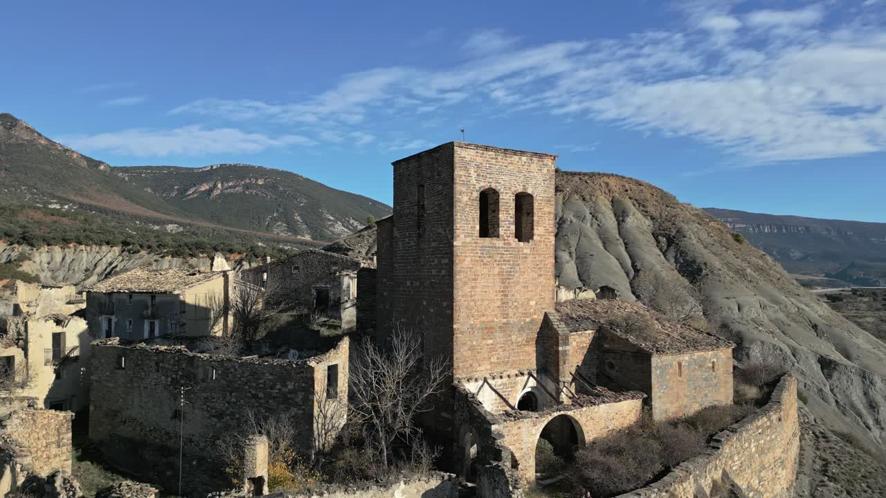 Filmato aereo con drone che circonda le rovine della città medievale abbandonata di Esco in Aragona, Spagna, rivelando la chiesa in pietra fatiscente e le case in rovina sullo sfondo di un paesaggio montano brullo.