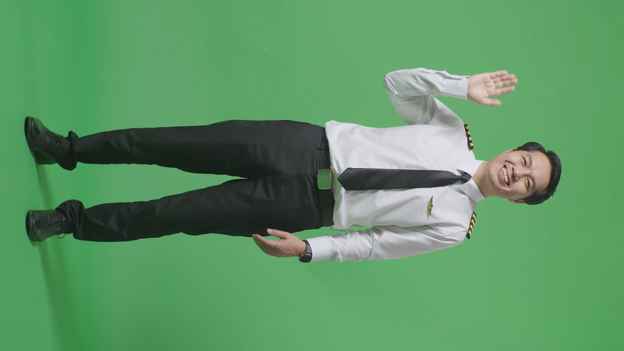 아시아 남성 조종사가 미소 짓고, 손을 흔들며, 녹색 스크린 배경 스튜디오에 서서 작별 인사를 하고 있습니다.